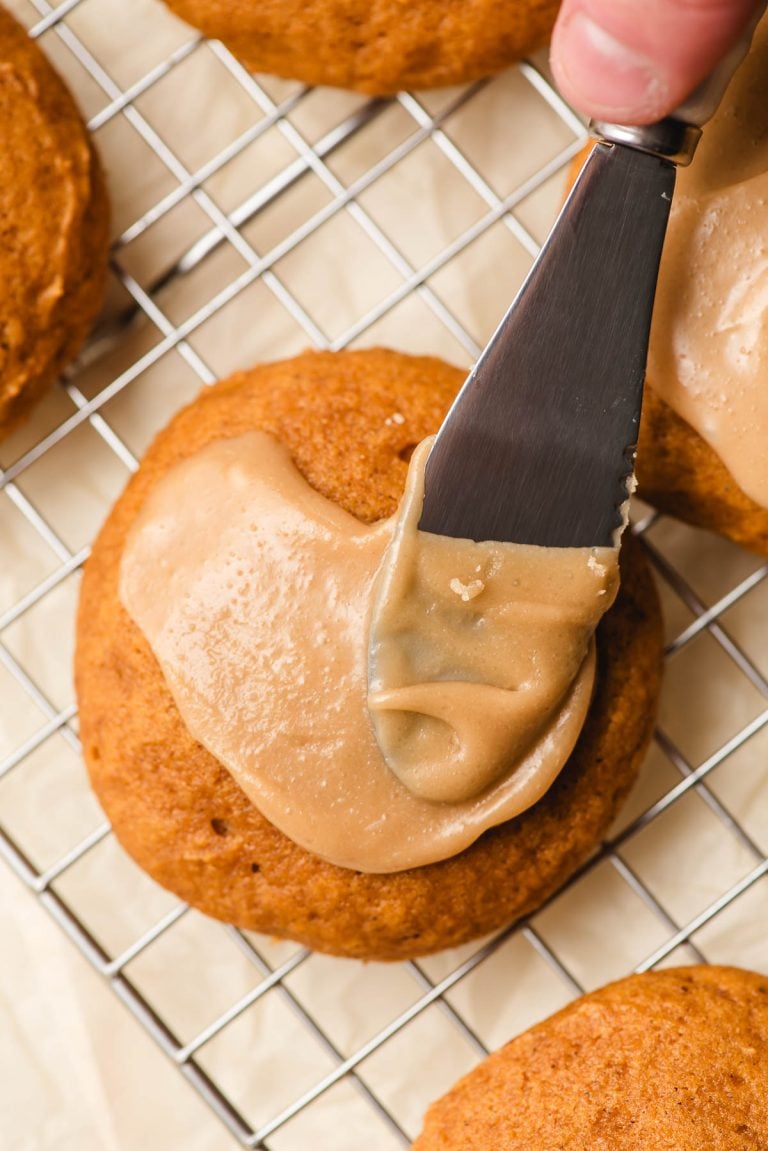 Spatula spreading penuche frosting onto a pumpkin cookie.