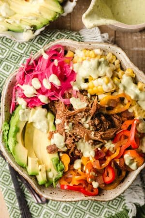 Scalloped bowl filled with rice, pulled pork, avocado, bell pepper, corn, and pickled cabbage and onions, drizzled with avocado dressing.