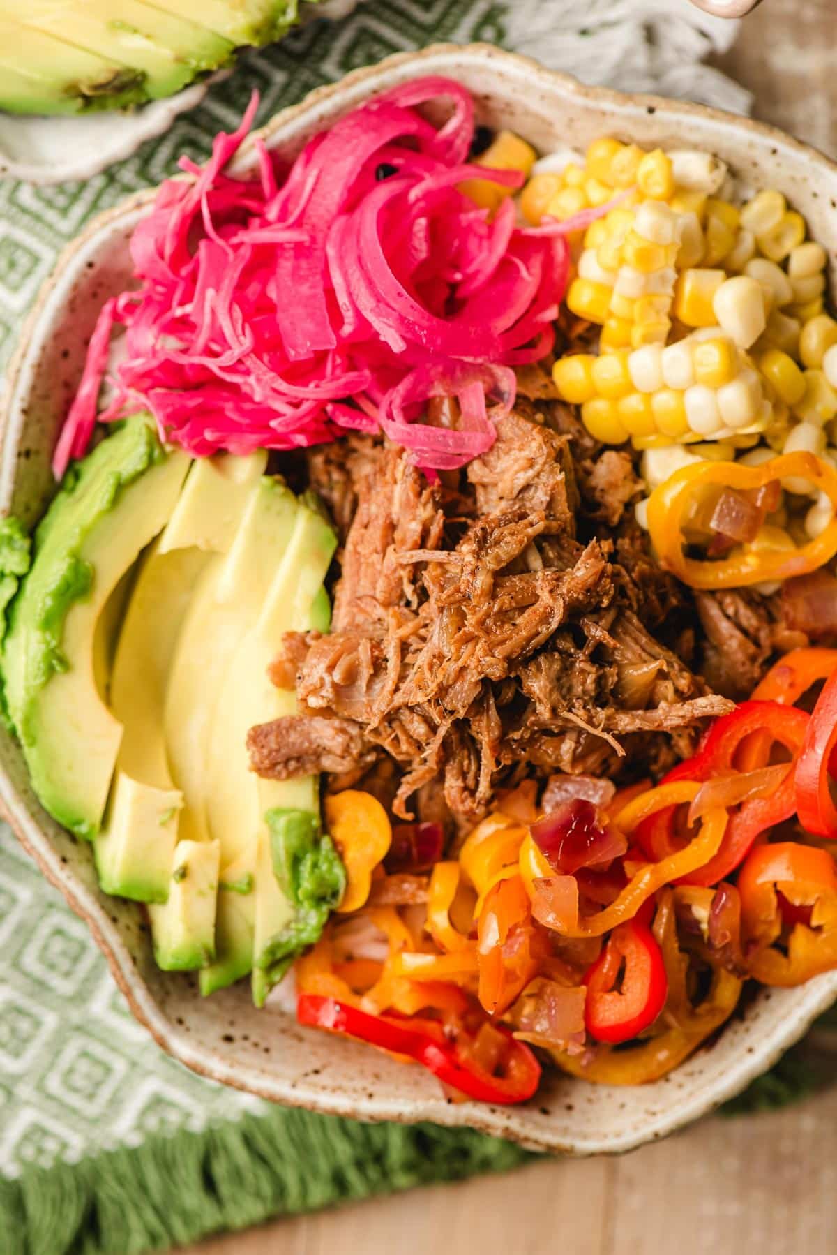 Rice bowl topped with pulled pork, avocado slices, sauteed peppers and onions, pickled onions, and corn.