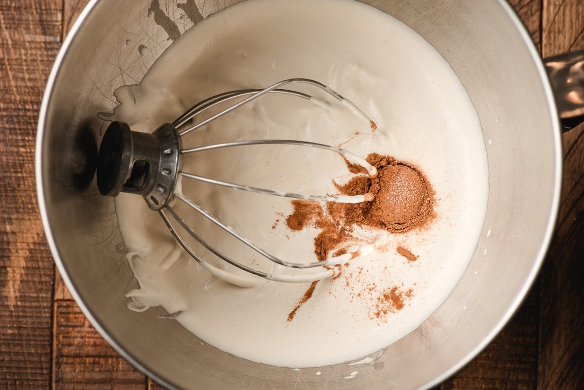 Cinnamon being added to lightly whipped cream in the bowl of an electric mixer.