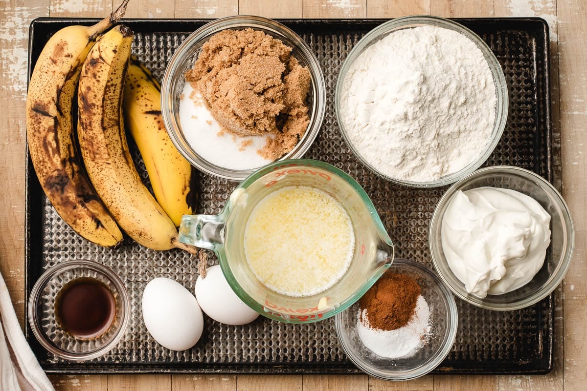 Bananas, brown sugar and white sugar, flour, baking soda, salt, cinnamon, melted butter, vanilla, and 2 eggs on a baking sheet.