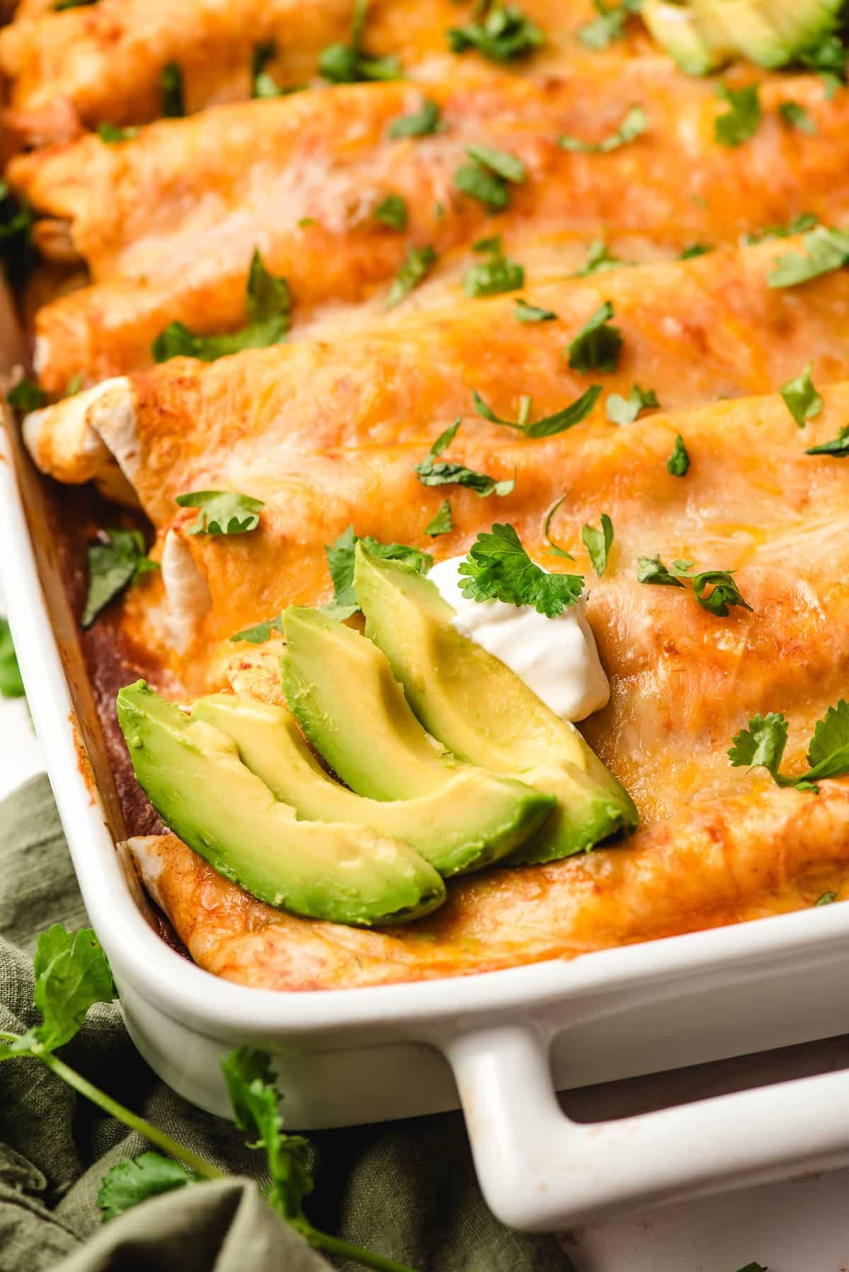 Casserole dish of cheesy chicken enchiladas topped with avocado slices.