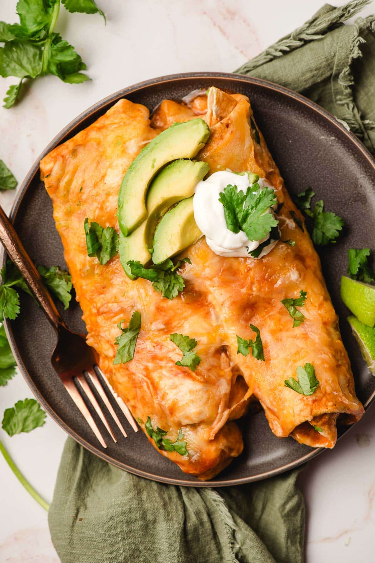 Two cheesy chicken enchiladas on a plate with avocado slices and sour cream.
