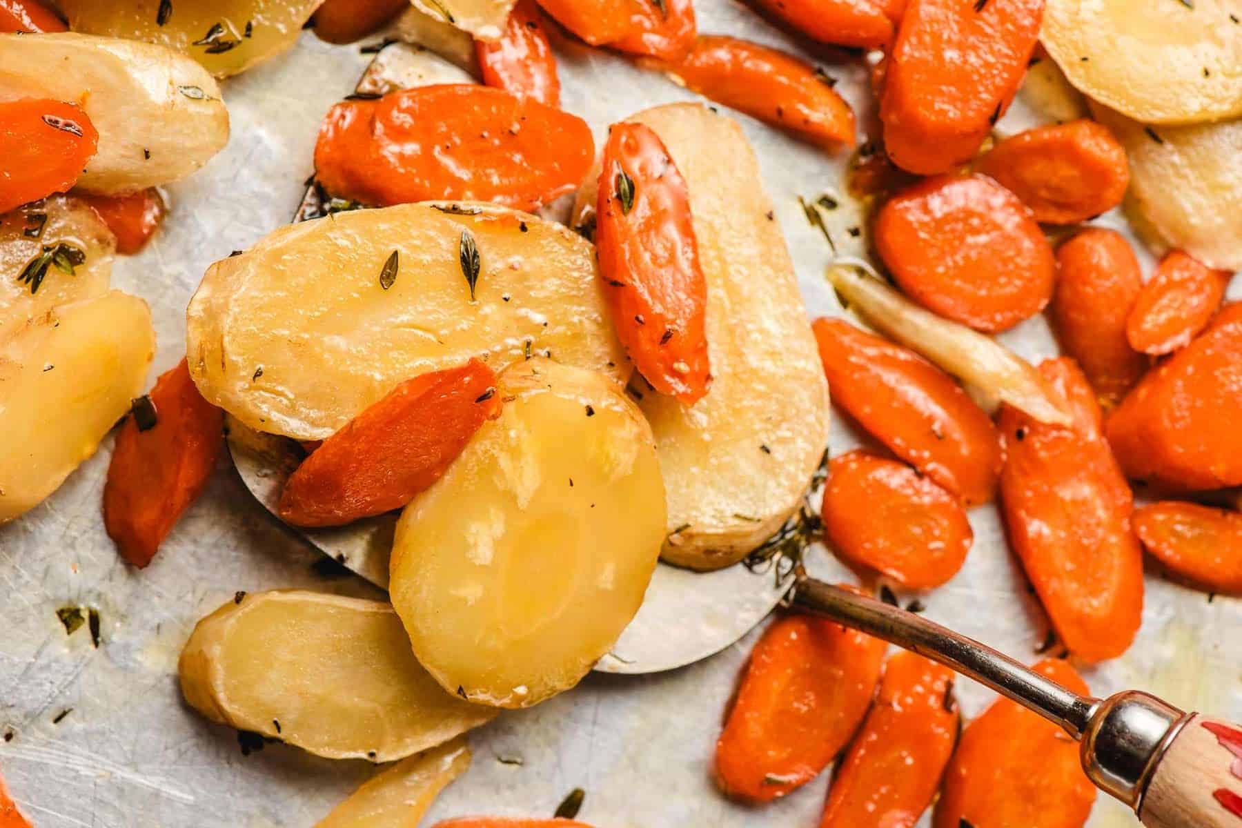 Metal spatula scooping up roasted carrots and parsnips from a sheet pan.