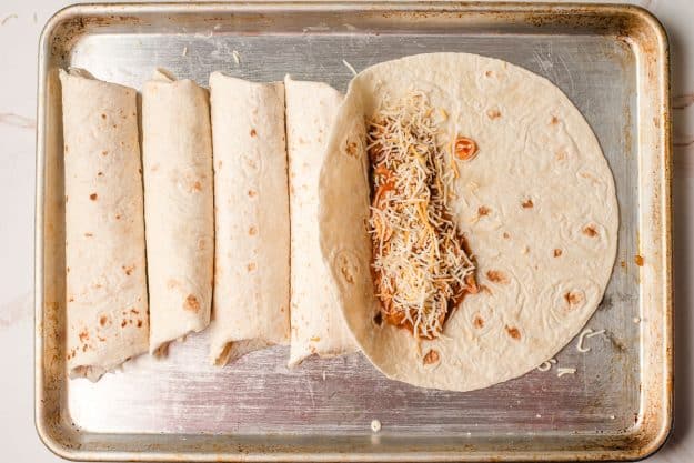 Chicken and cheese being loaded and rolled into tortillas on a baking sheet.