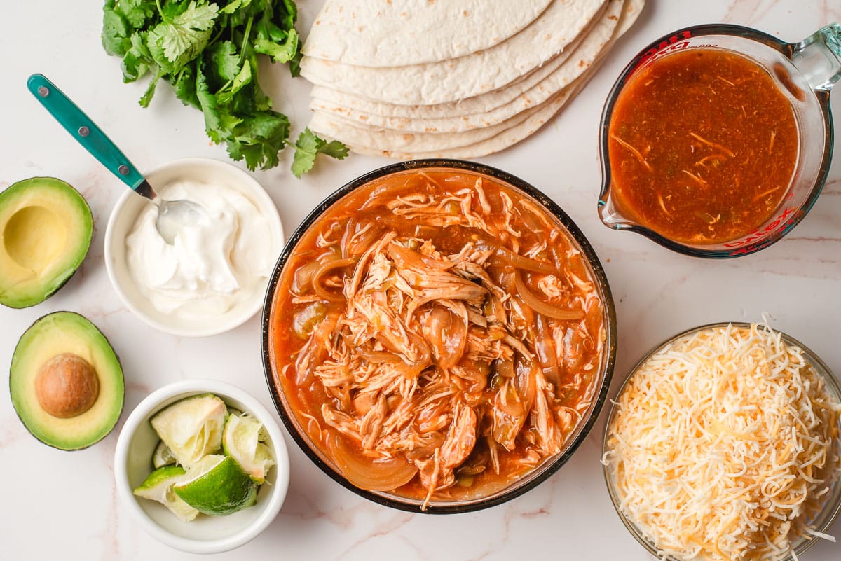 Shredded enchilada chicken, enchilada sauce, avocado, cheese, tortillas, cilantro, sour cream, and lime wedges on a white background.