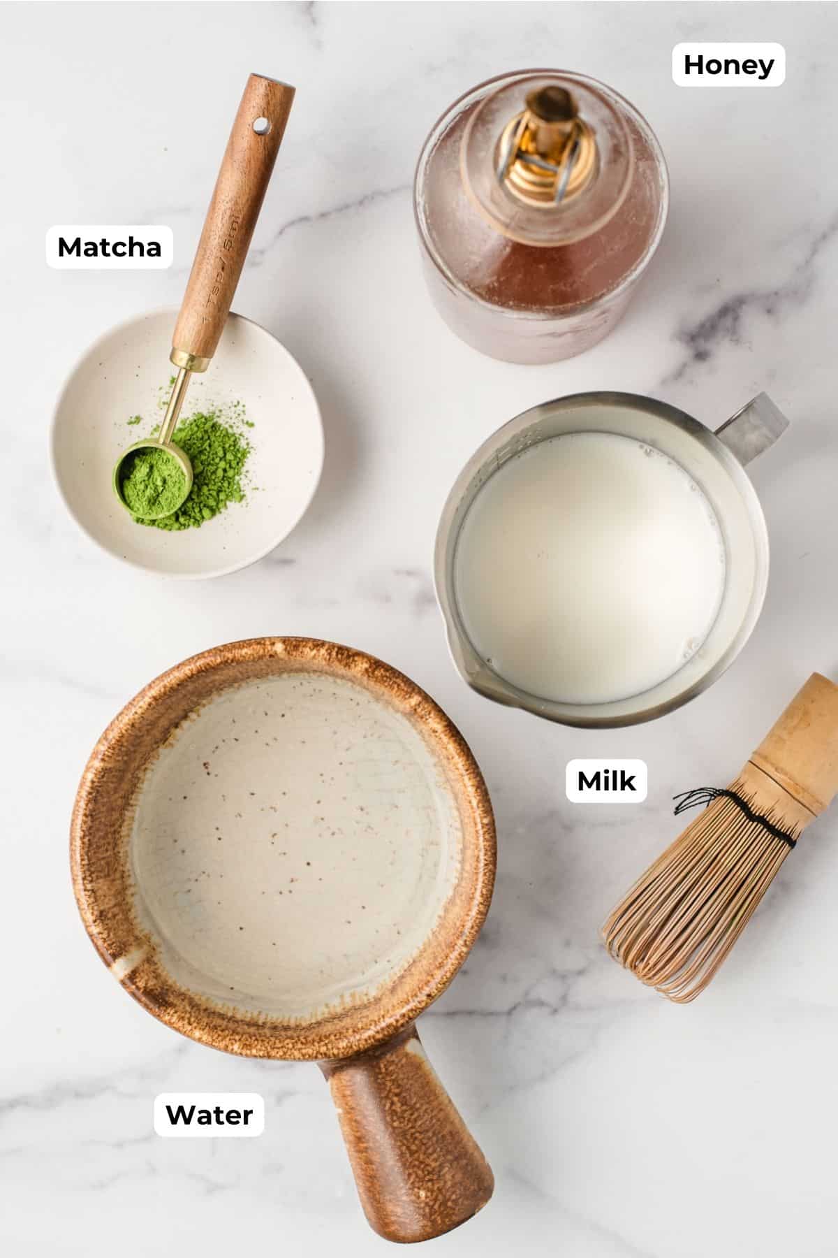 recipe ingredients for a hot matcha latte including match powder, milk, a bamboo whisk, and a sifter on a kitchen counter. 