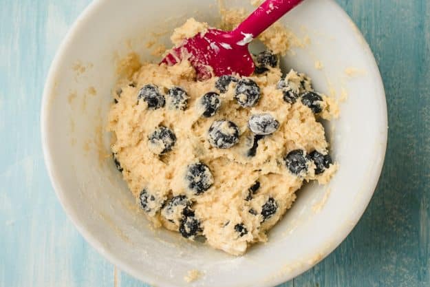 Fresh blueberries being stirred into coffee cake batter in a white bowl.