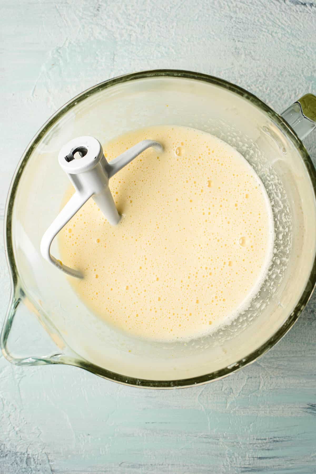 A glass mixing bowl contains smooth, pale yellow batter with a paddle attachment resting in it. The bowl is placed on a light blue textured surface.