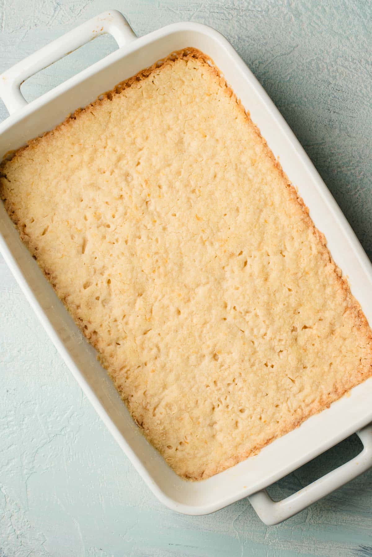 Baked shortbread crust in a rectangular white dish on a textured light blue surface. The crust is golden brown with slightly uneven edges.