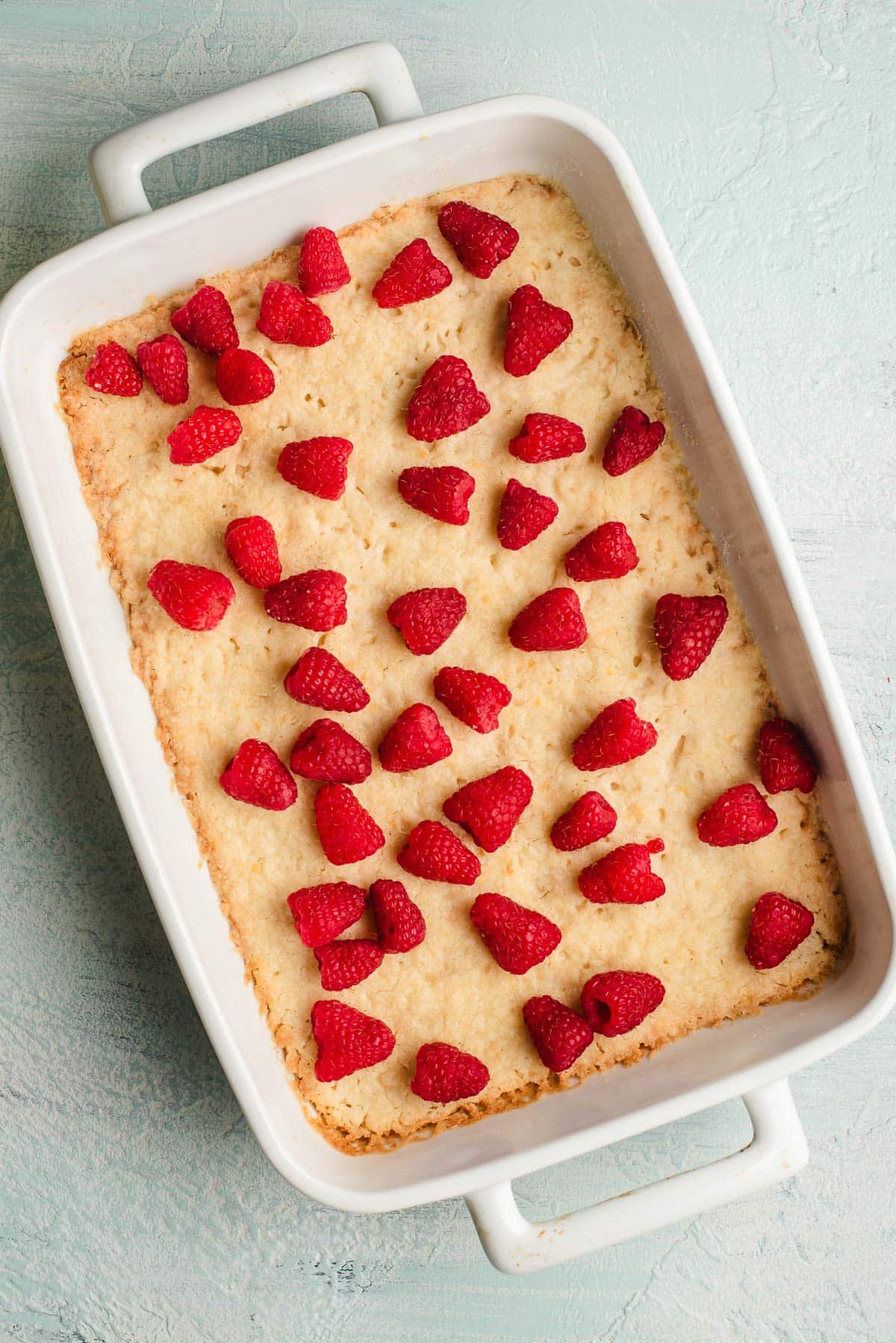 A rectangular white baking dish filled with a creamy baked crust is topped with fresh raspberries evenly scattered on top. The dish is set on a light textured surface.