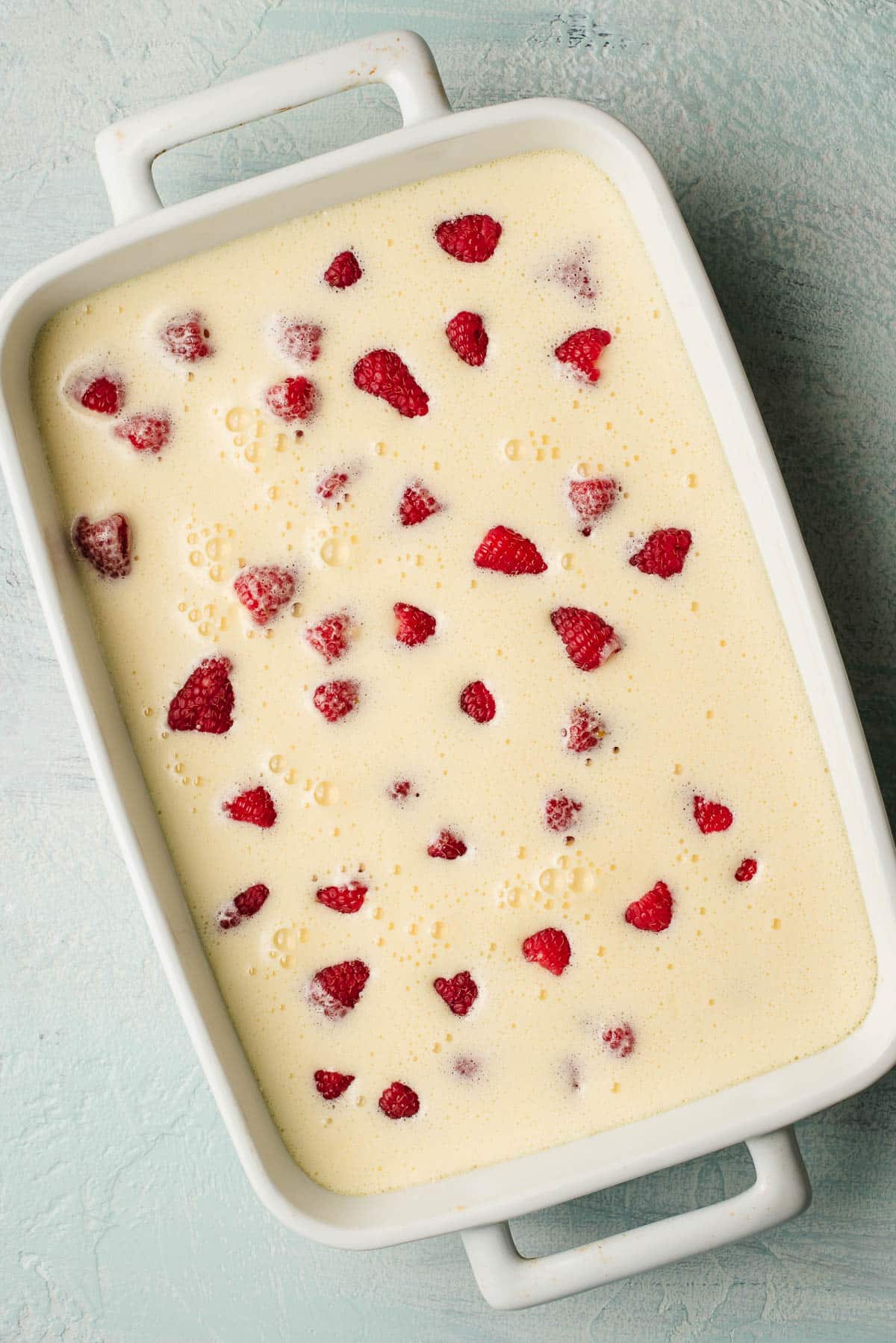 A rectangular baking dish filled with a creamy custard mixture is topped with fresh raspberries, ready for baking. The dish has handles and sits on a textured light blue surface.