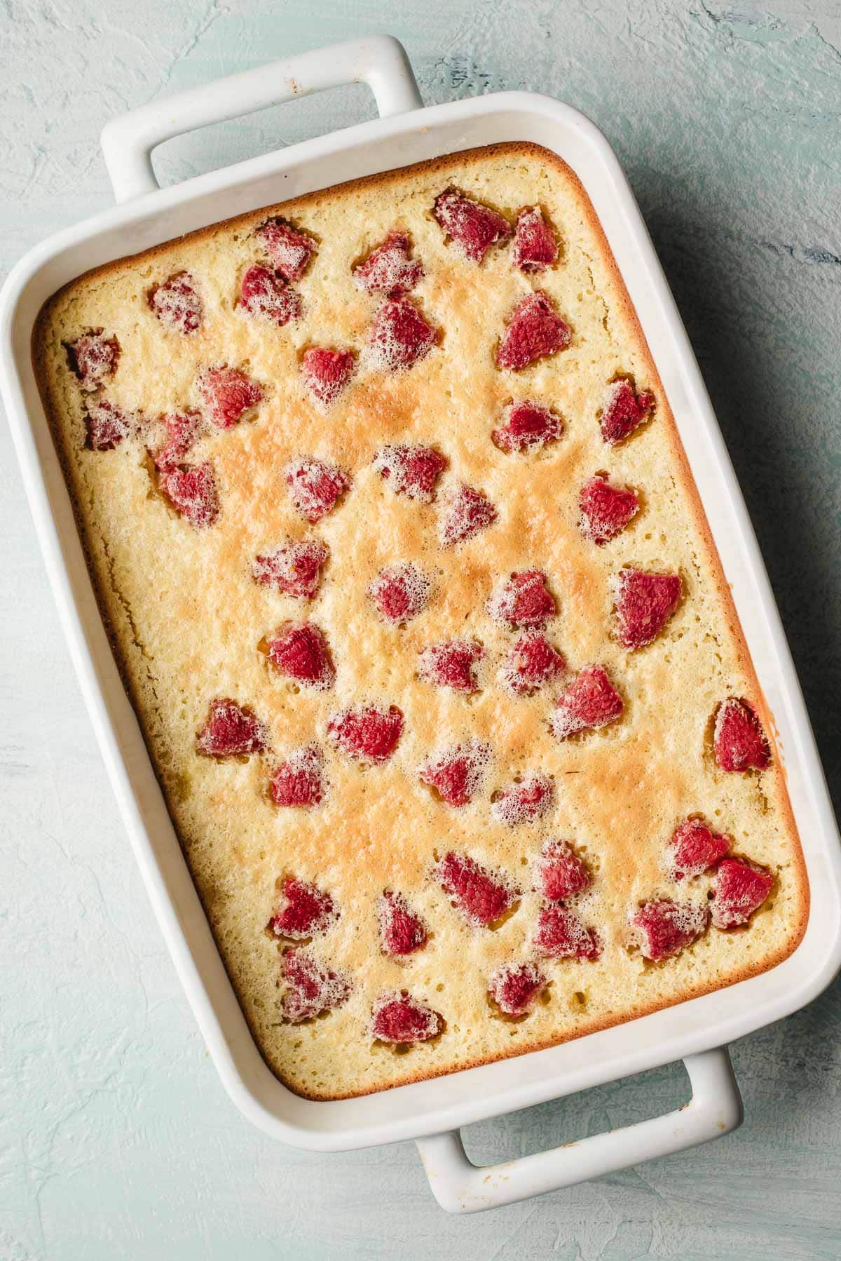 baked lemon raspberry bars in a rectangular white dish, topped with scattered pieces of red fruit on a golden-brown surface, sits on a light-textured surface.