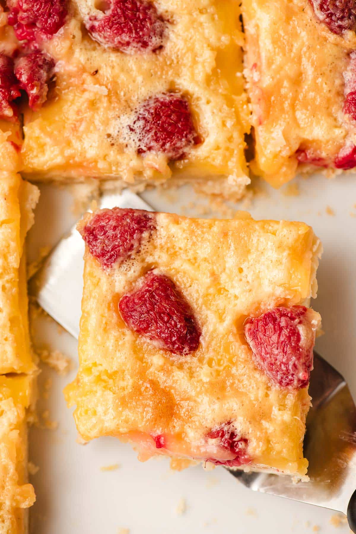 A close-up of a slice of a lemon raspberry bar being lifted by a spatula. Other slices are visible nearby.