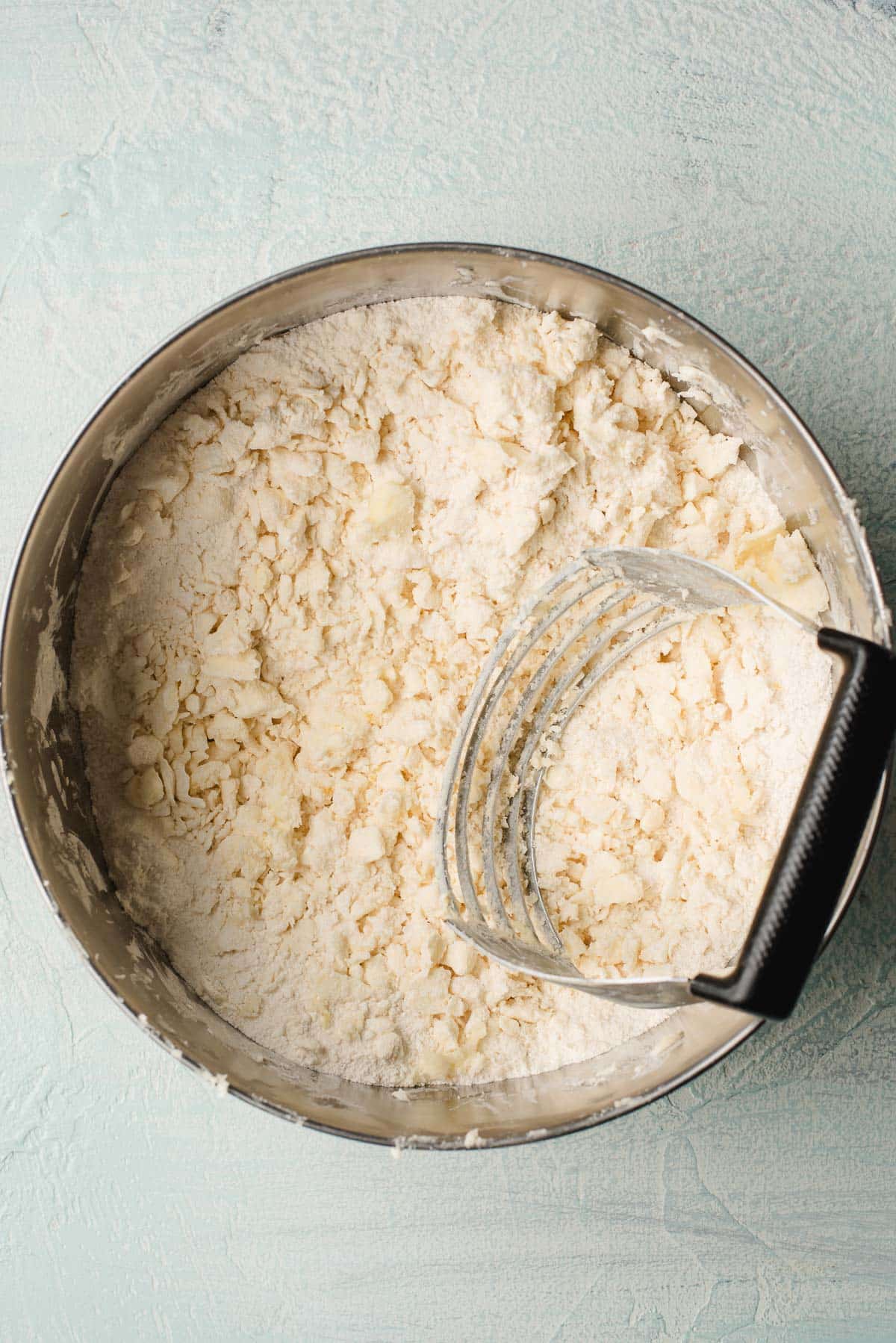 A metal bowl filled with a mixture of flour and butter, being cut with a pastry cutter. The texture is coarse, resembling crumbs, against a light blue background.