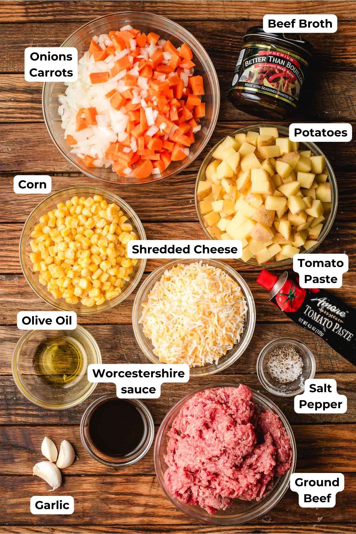 An assortment of ingredients for a ground beef and potatoes on a wooden surface, including chopped onions, carrots, potatoes, corn, shredded cheese, beef broth, tomato paste, olive oil, Worcestershire sauce, garlic cloves, salt, pepper, and ground beef.