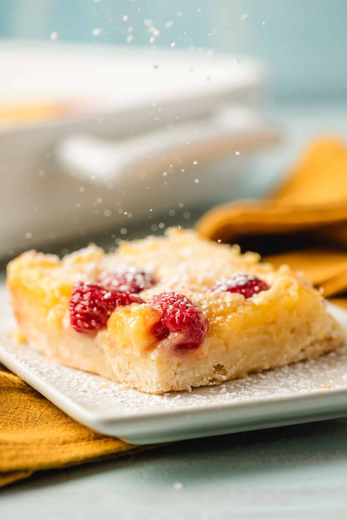 A slice of raspberry lemon bars with a crumbly base topped with powdered sugar and fresh raspberries sits on a white plate. In the background, a yellow napkin and a baking dish are slightly blurred.