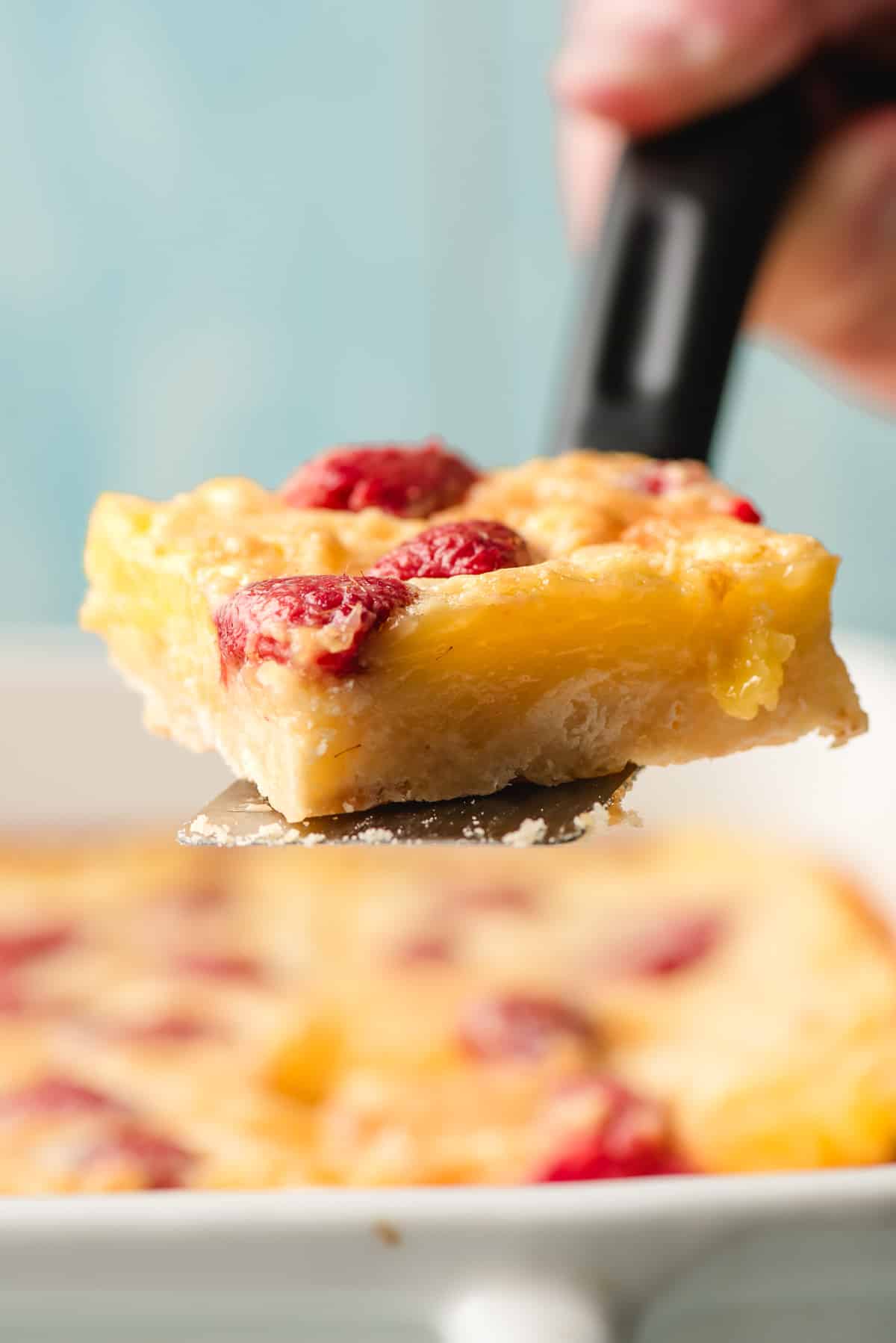 A slice of lemon raspberry bars with a golden crust and raspberry pieces being lifted from a baking dish with a spatula. The background shows the rest of the dessert in the dish.
