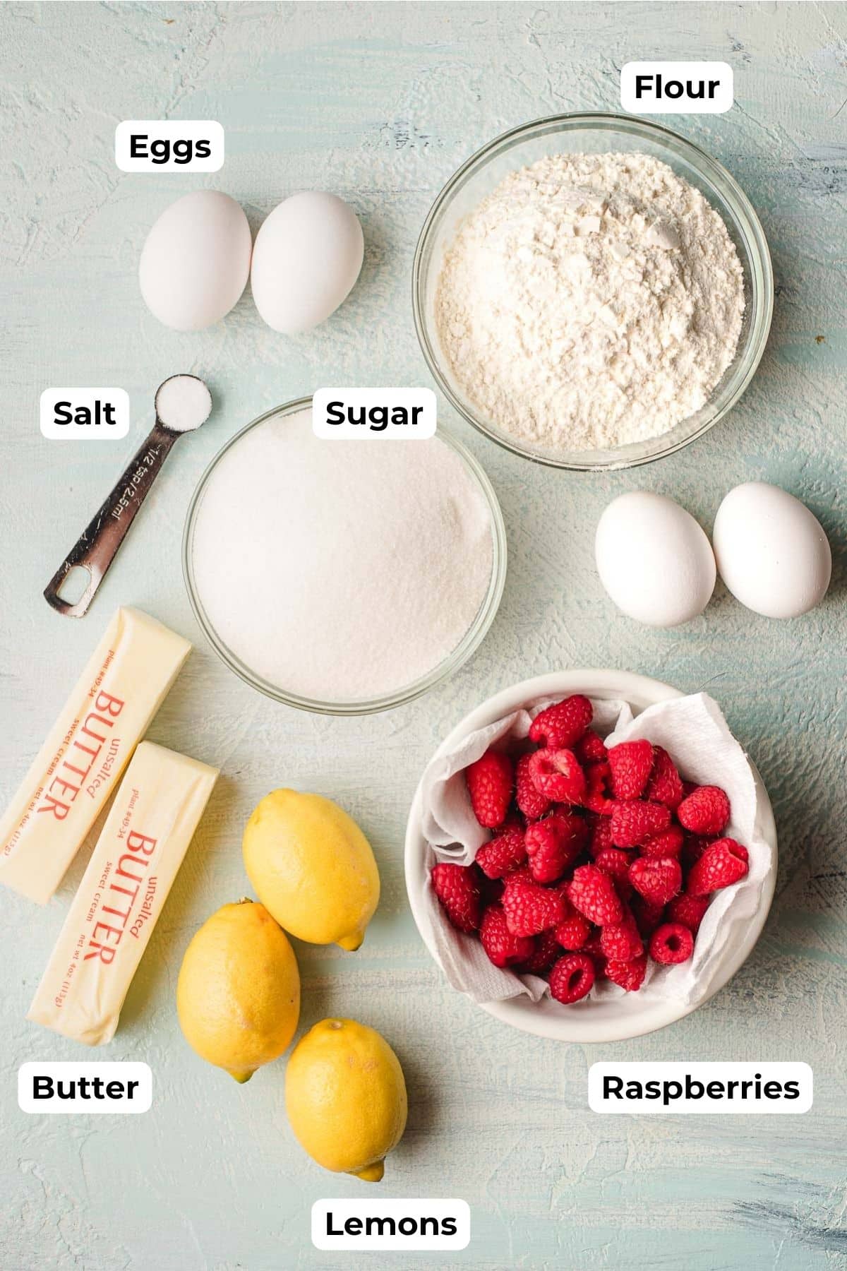 Ingredients for lemon raspberry bars displayed on a surface: eggs, flour in a bowl, salt in a measuring spoon, sugar in a bowl, butter sticks, fresh raspberries in a bowl, and whole lemons. Each item is labeled.
