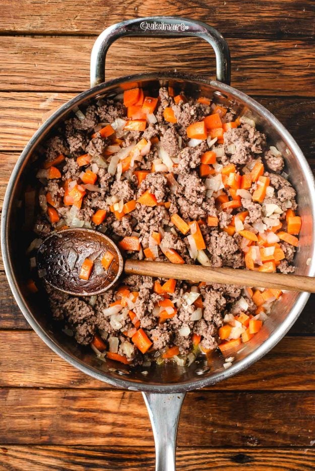 A skillet filled with cooked ground beef, chopped carrots, and onions on a wooden surface. A wooden spoon rests in the pan, stirring the ingredients.