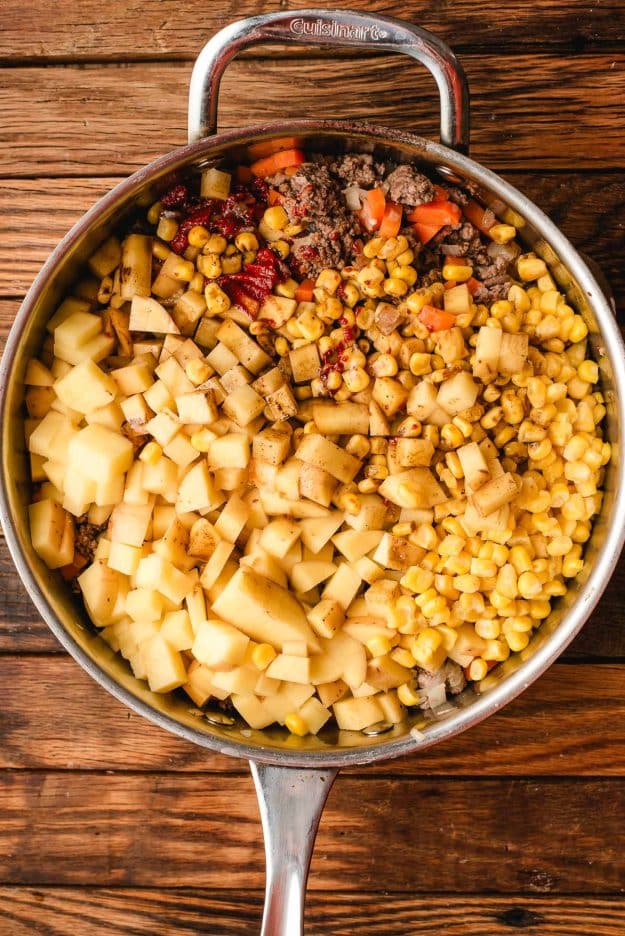 A skillet on a wooden surface filled with browned ground beef, diced potatoes, corn kernels, chopped carrots, and chopped red bell peppers, all evenly distributed without being mixed.