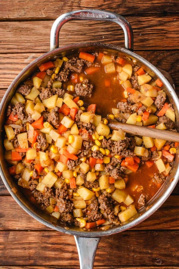 ground beef and potatoes skillet on a wooden surface, containing ground beef, diced potatoes, carrots, corn, and red bell peppers in a savory broth. A wooden spoon rests inside the pot.