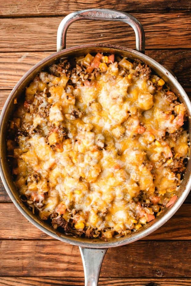 A skillet filled melted cheese, ground beef, diced vegetables, and corn. The skillet has a shiny metal handle and is set on a wooden surface.