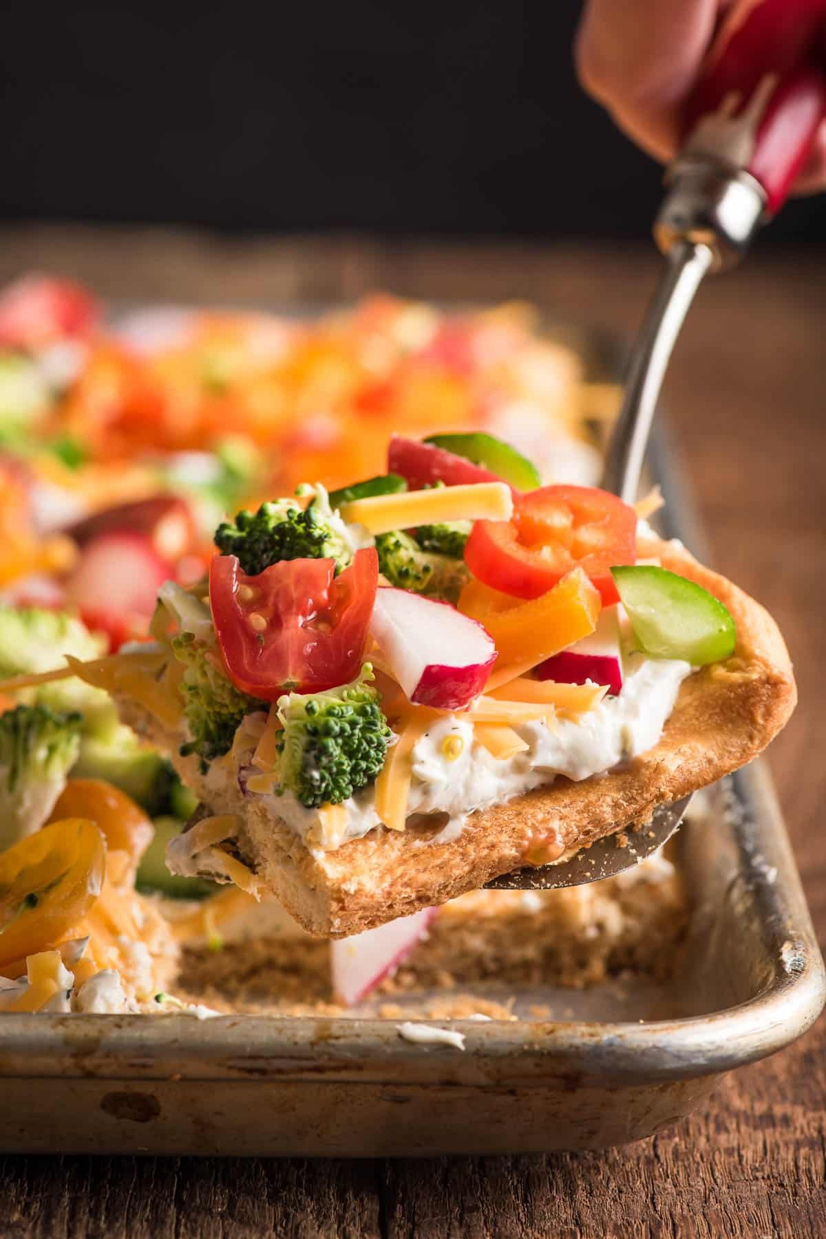 Close-up of cold veggie pizza slice topped with fresh raw vegetables and shredded cheese.
