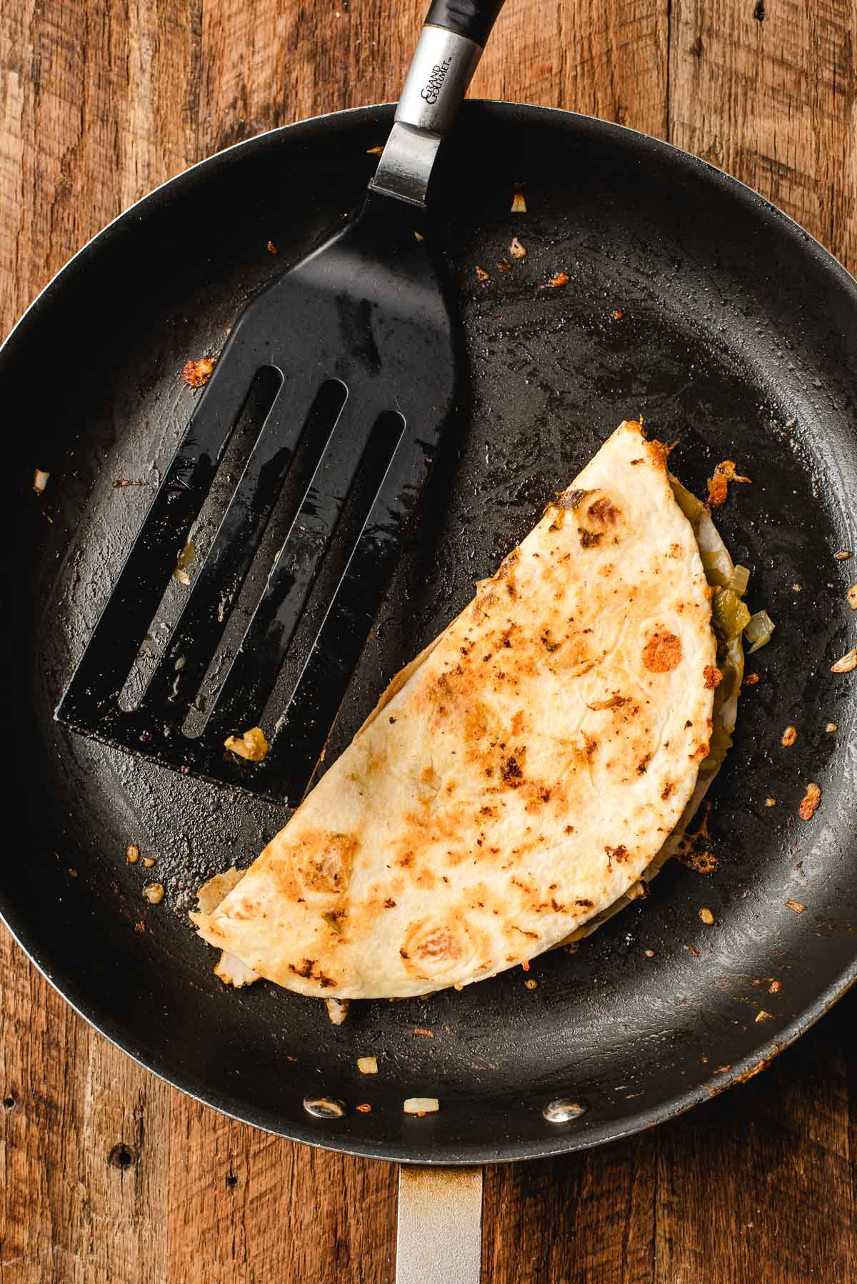 Crispy folded quesadilla in skillet being pressed with a spatula.