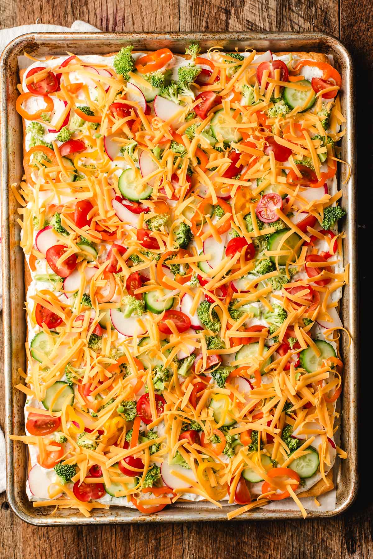 Chopped vegetables and shredded cheddar being sprinkled over cream cheese layer.