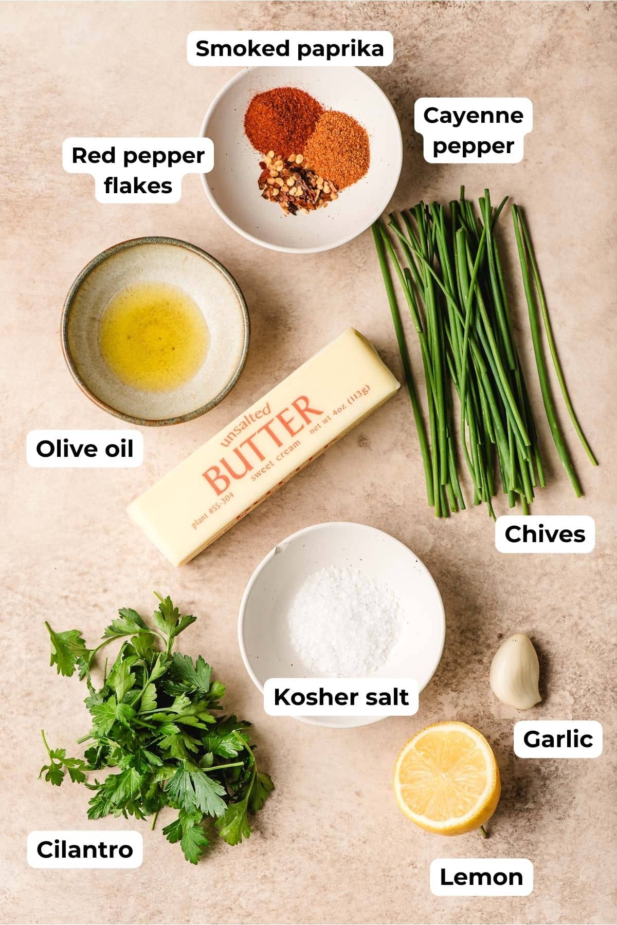 Stick of butter surrounded by olive oil, bowls of spices, parsley, chives, halved lemon, and garlic clove.