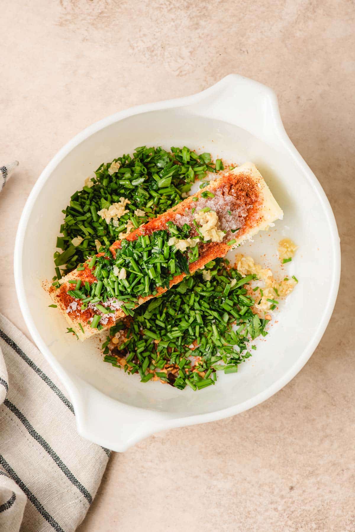 Stick of butter in a white bowl, topped with chopped parsley and chives, garlic, and spices.