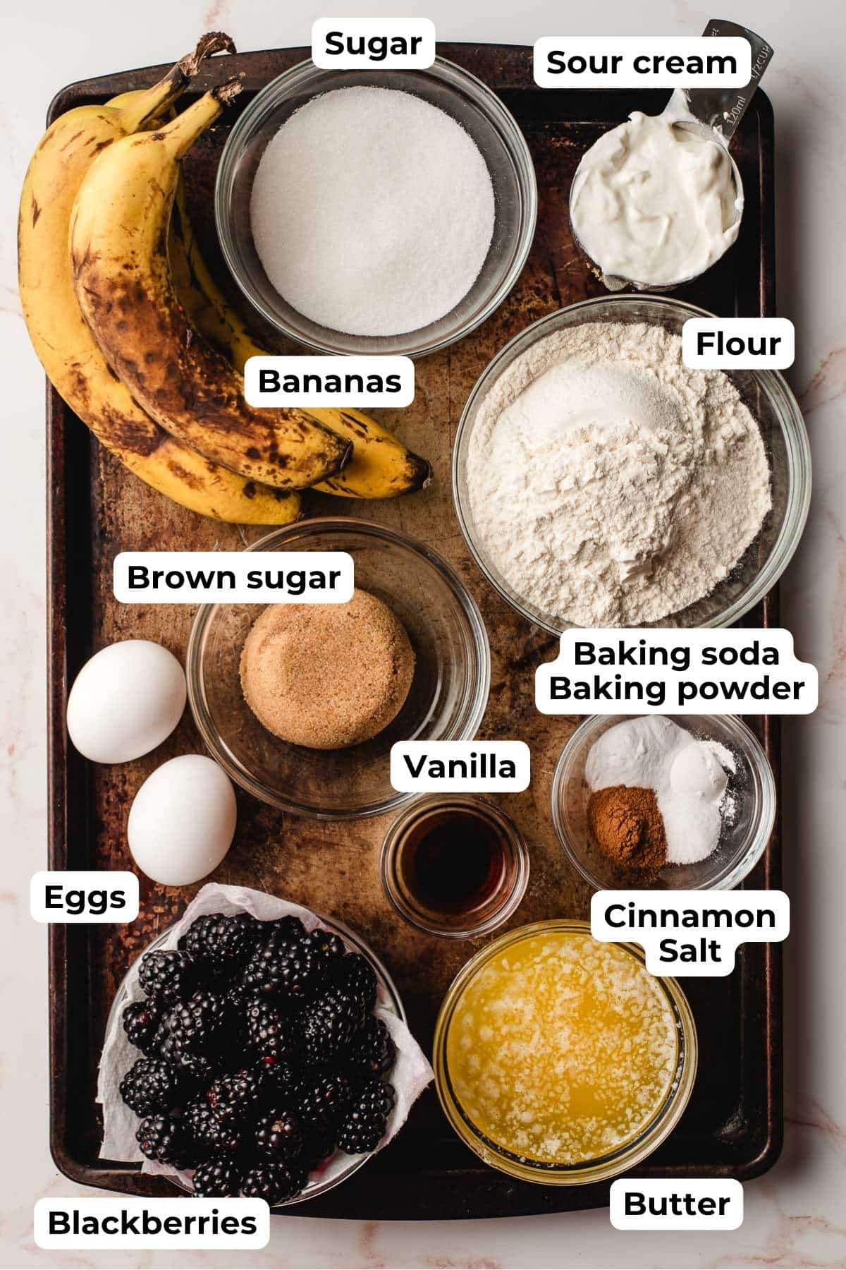 ingredients for banana blackberry muffins in small bowls on a baking sheet including bananas, butter, vanilla, salt, cinnamon, flour, sour cream, baking powder, baking soda, brown sugar, blackberries, and and egg