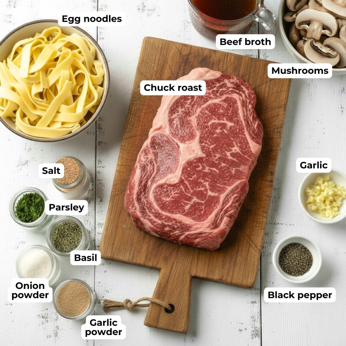 Chuck roast on a wooden cutting board surrounded by jars of spices, a bowl of egg noodles, mushrooms, beef broth, and minced garlic.