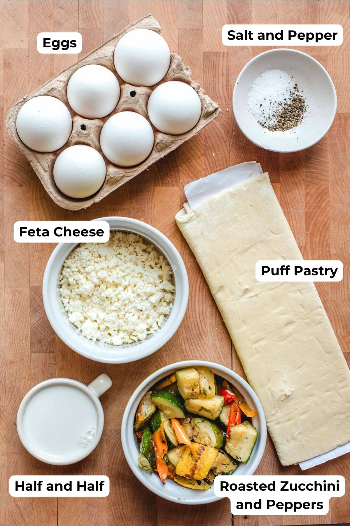 Eggs, salt and pepper, feta cheese, puff pastry, half and half, and roasted vegetables on a cutting board.