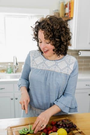 White woman with curly hair and a blue shirt chopping fruit on a cutting board.