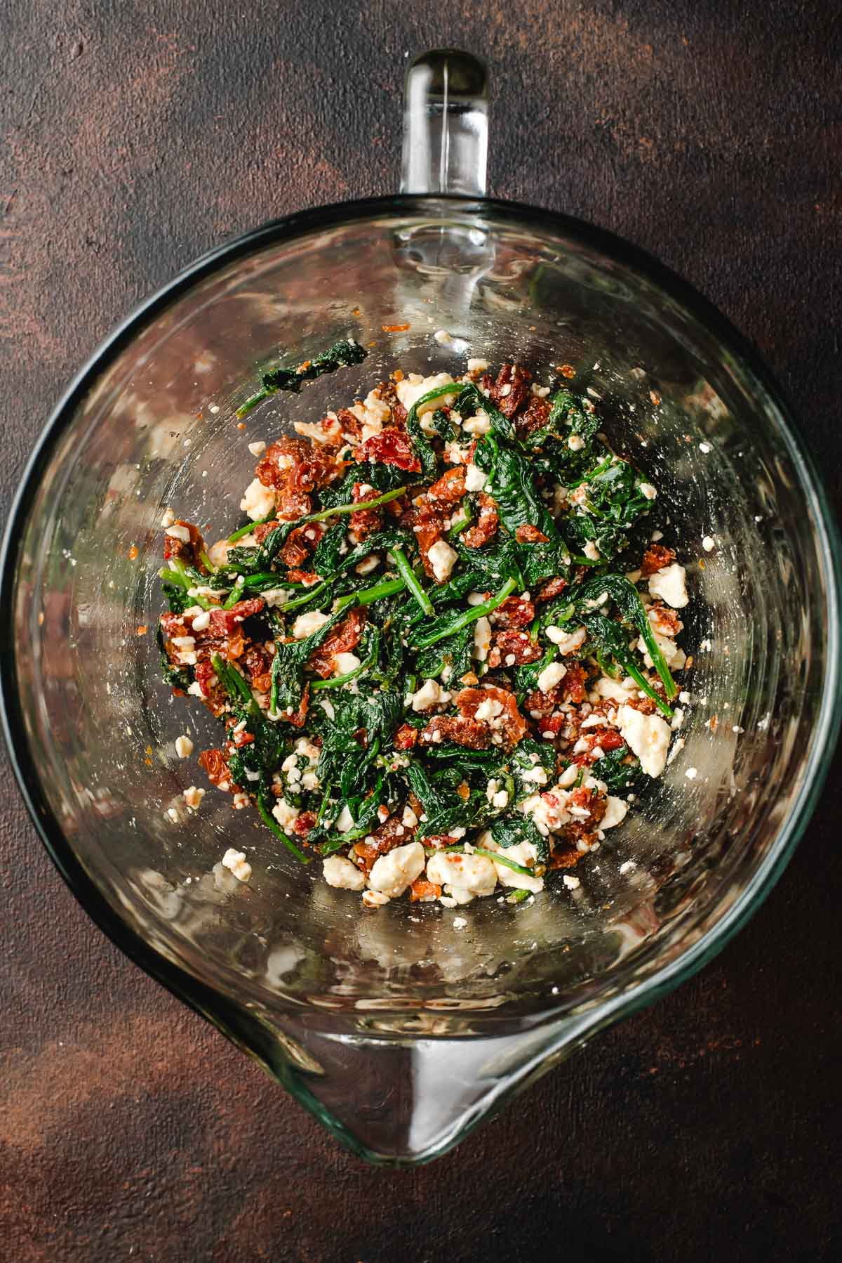 feta, sun-dried tomatoes, and cooked spinach in a glass measuring jar.