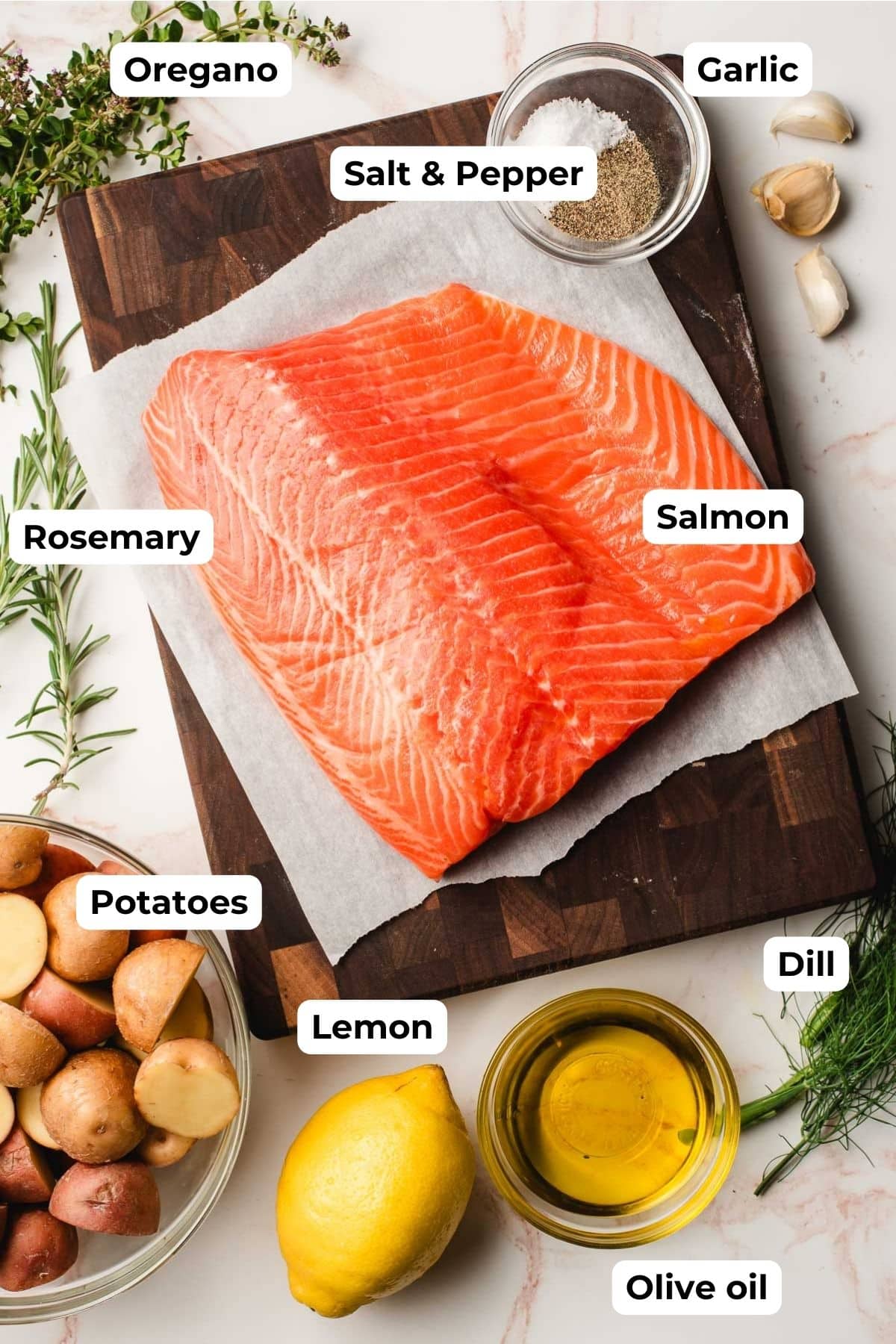Salmon fillet, baby potatoes, lemon, olive oil, dill, rosemary, oregano, garlic cloves, and salt and pepper on a white background.