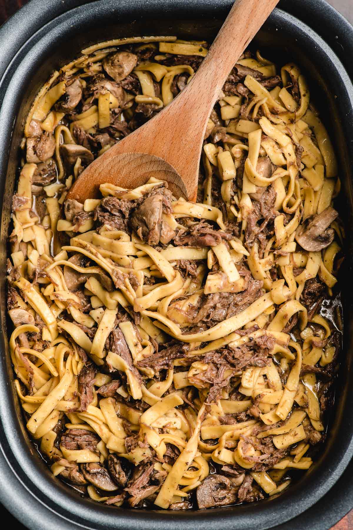 shredded beef and egg noodles in a crock pot, getting mixed with a wooden spoon. 