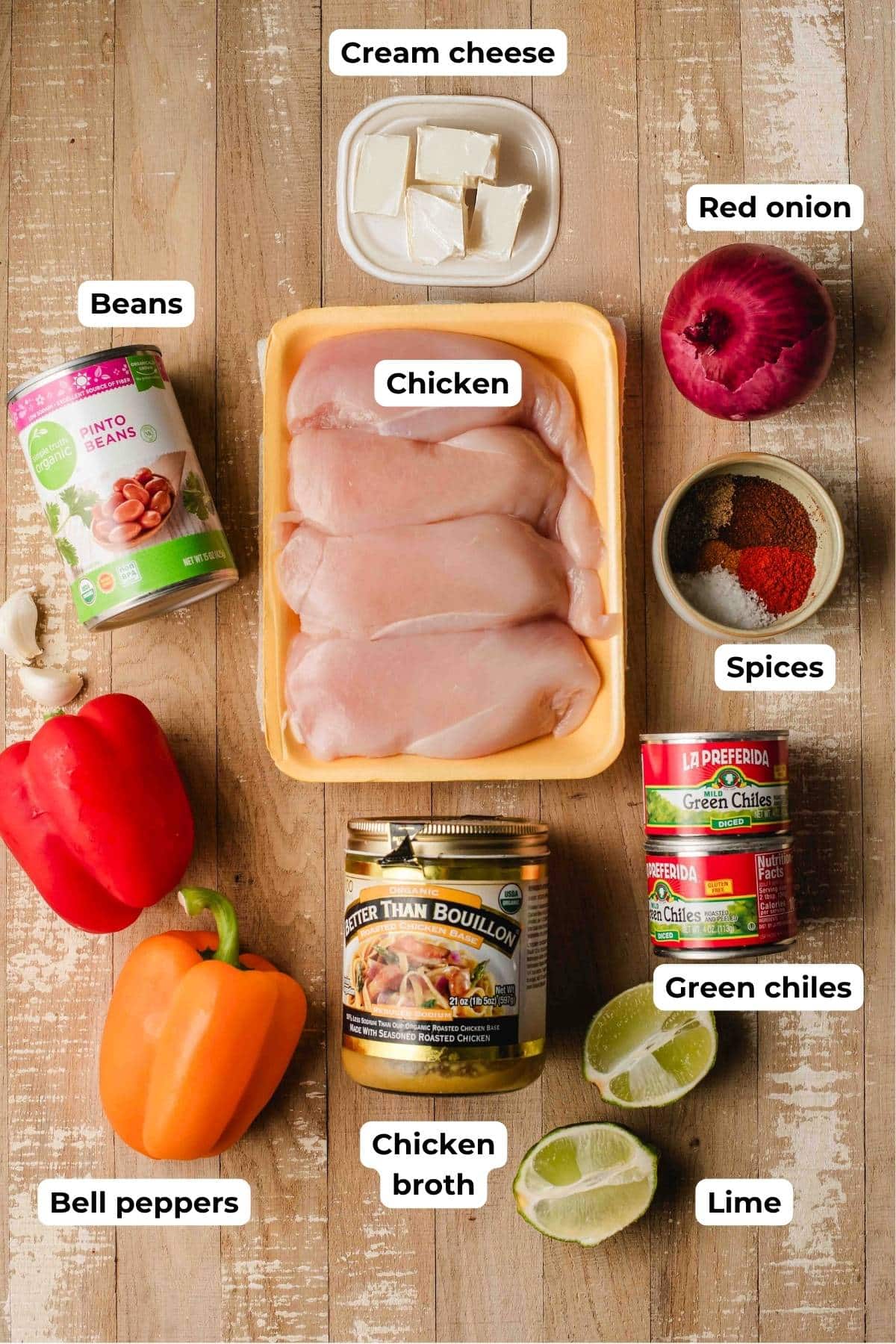 Chicken, beans, cream cheese, red onion, spices, green chiles, lime, better than bouillon, and bell peppers on a light wood background.