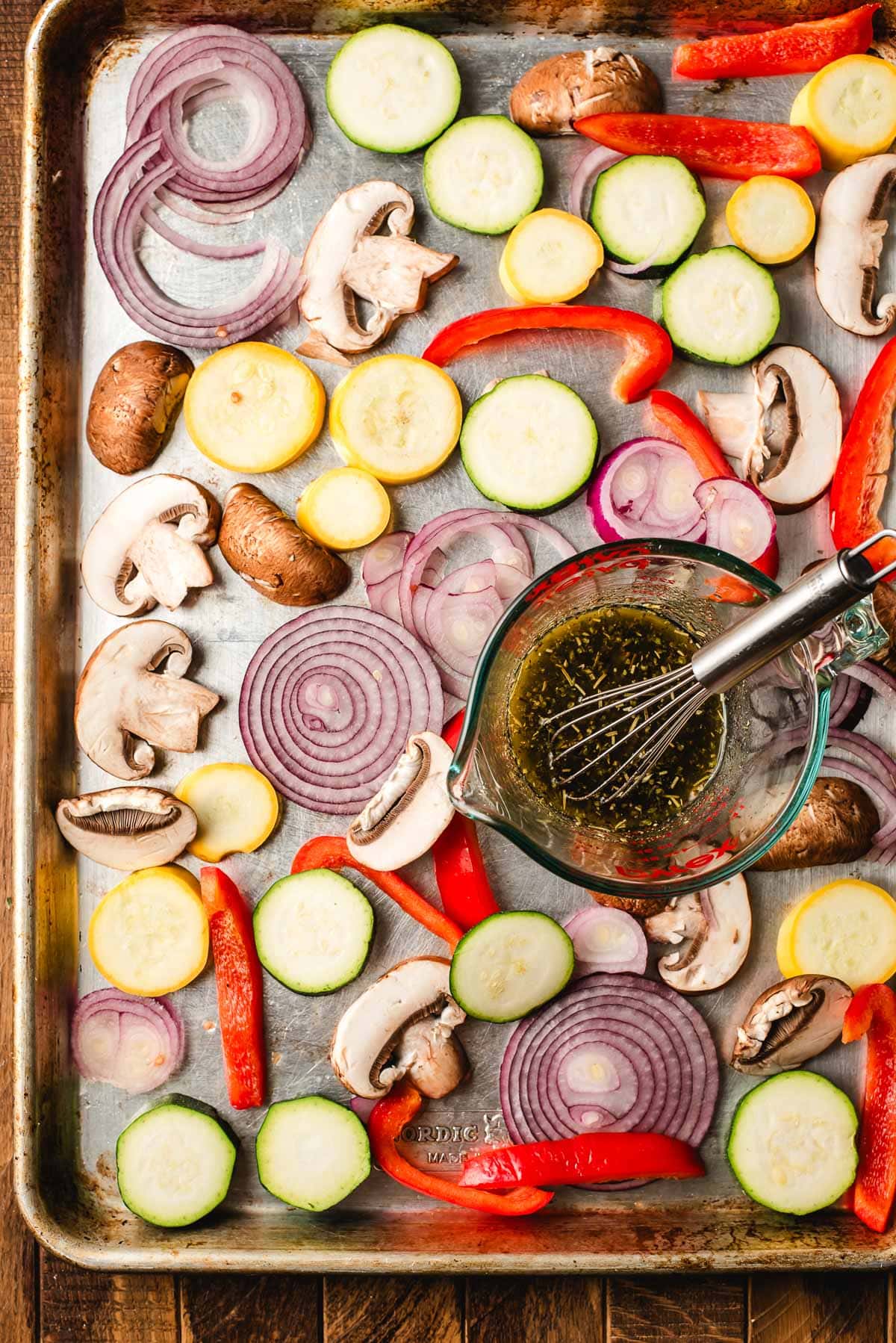 Sliced veggies on a sheet pan with a glass measuring cup filled with olive oil, red wine vinegar, and seasonings.