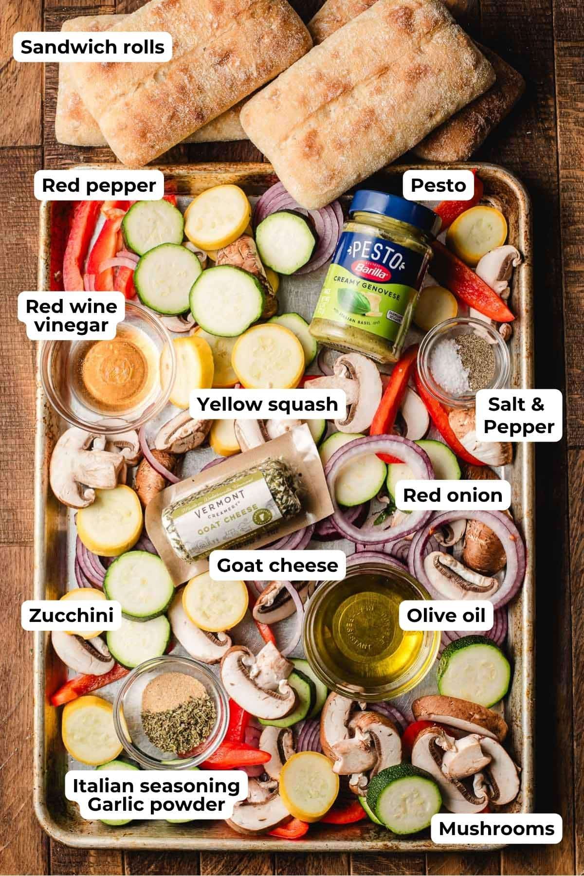 Sandwich rolls, chopped vegetables, seasonings, red wine vinegar, olive oil, pesto, and goat cheese arranged on a baking sheet.