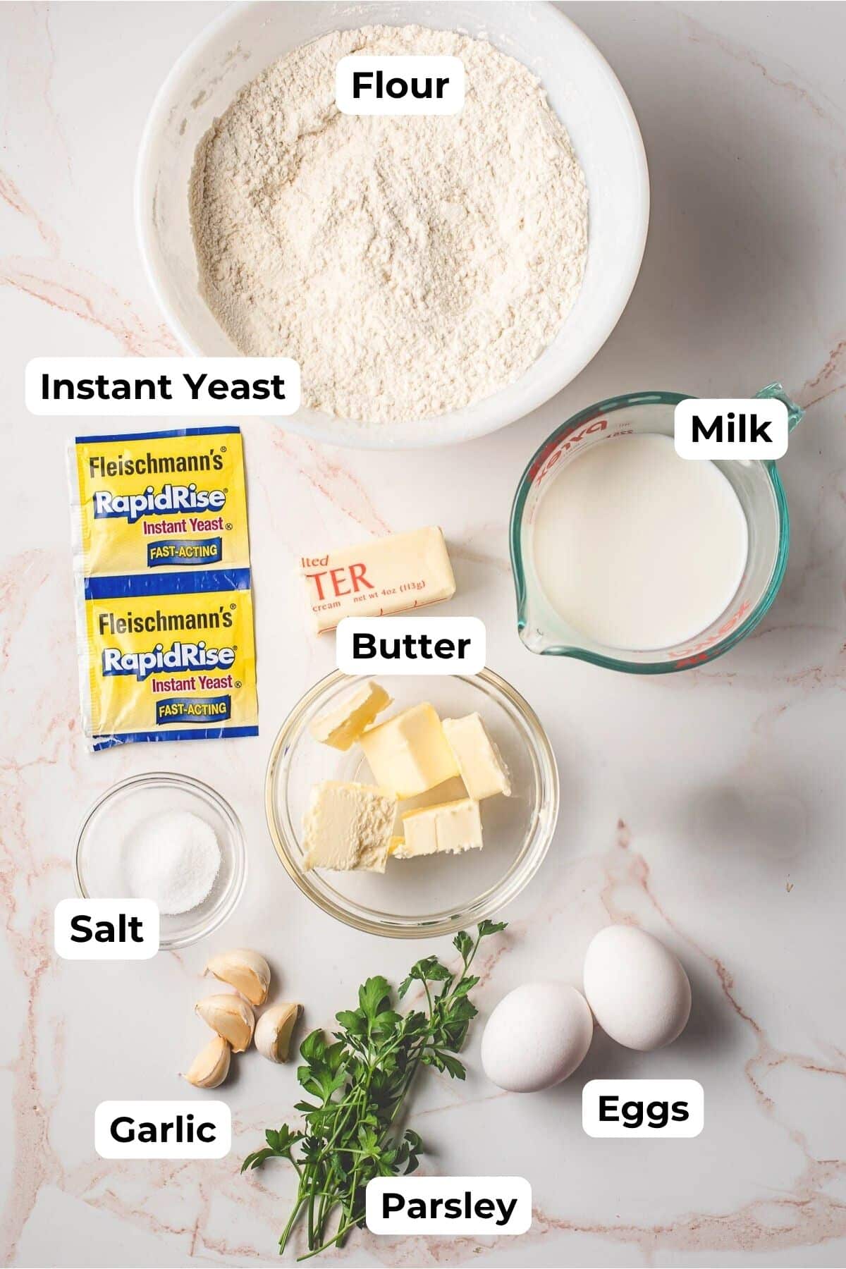 Flour, yeast, butter, milk, salt, parsley, garlic, and eggs on a white marble background.