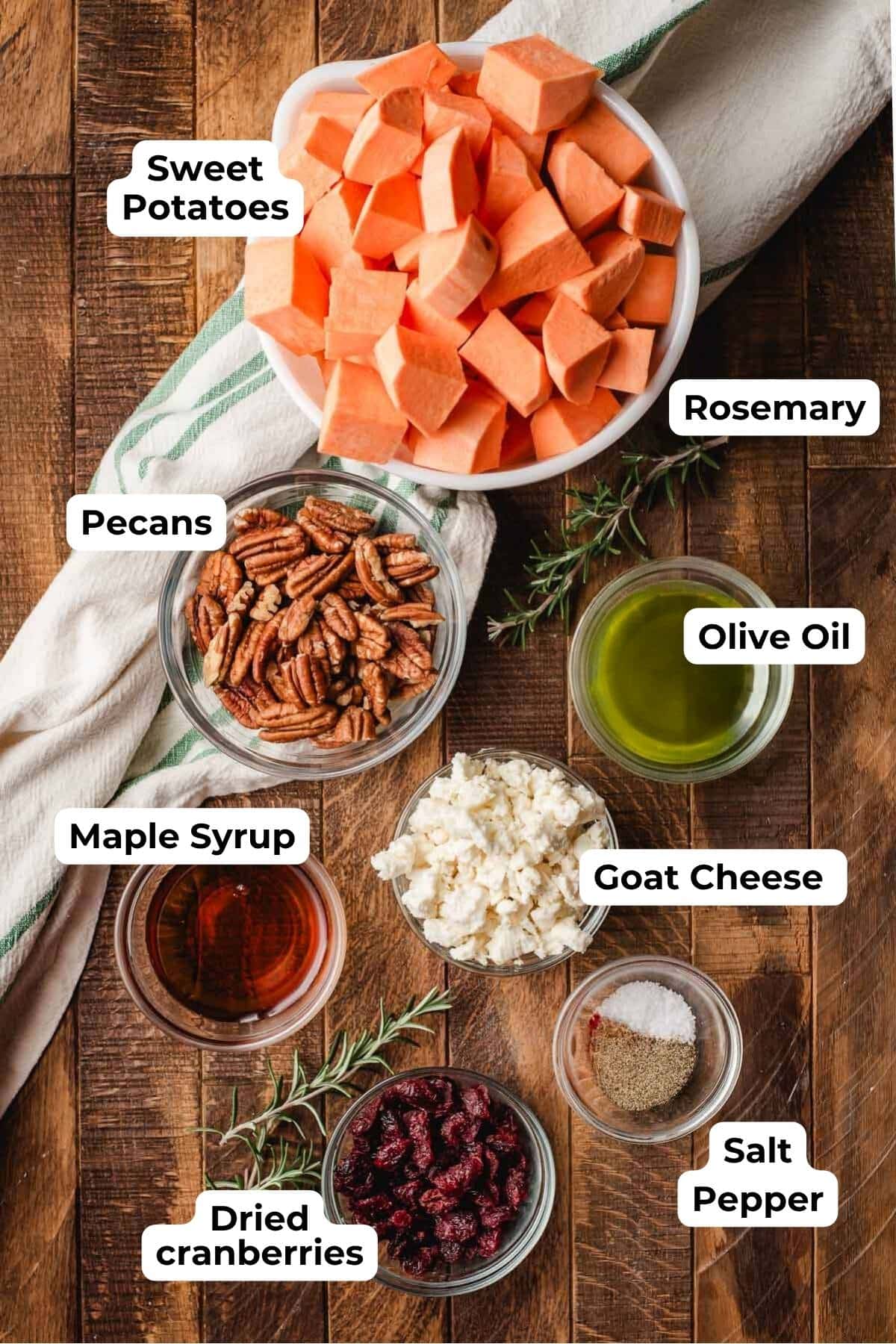 Chopped sweet potatoes, pecans, maple syrup, goat cheese, olive oil, salt and pepper, and cranberries all in prep bowls on a wood background.