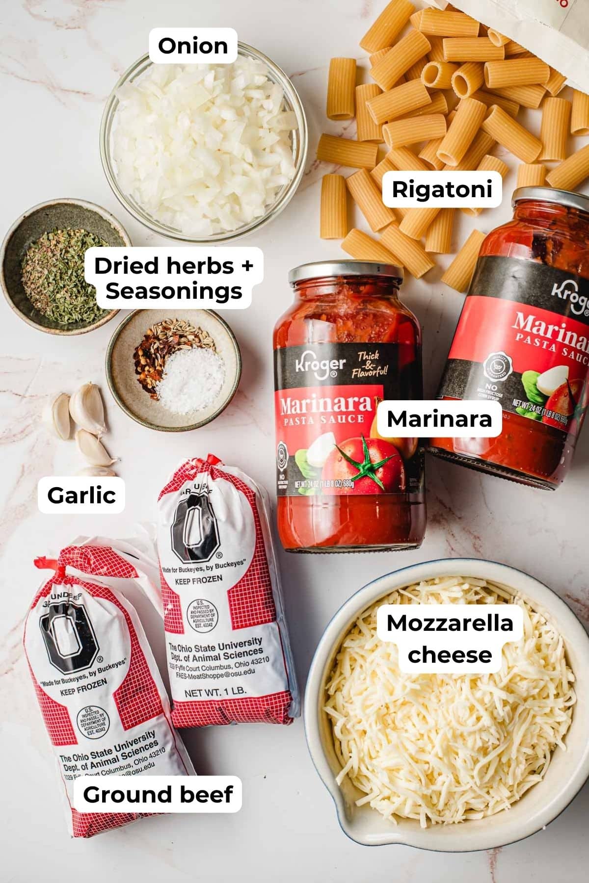 Ground beef, mozzarella cheese, seasonings, chopped onion, rigatoni, and marinara jars on a white background.