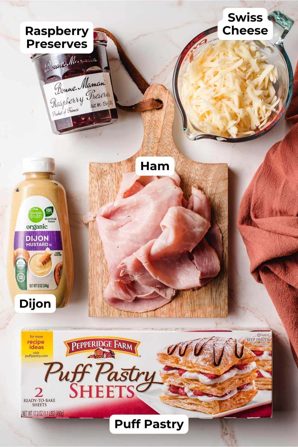 Ham, shredded Swiss cheese, dijon, puff pastry, and raspberry preserves on a marble background.