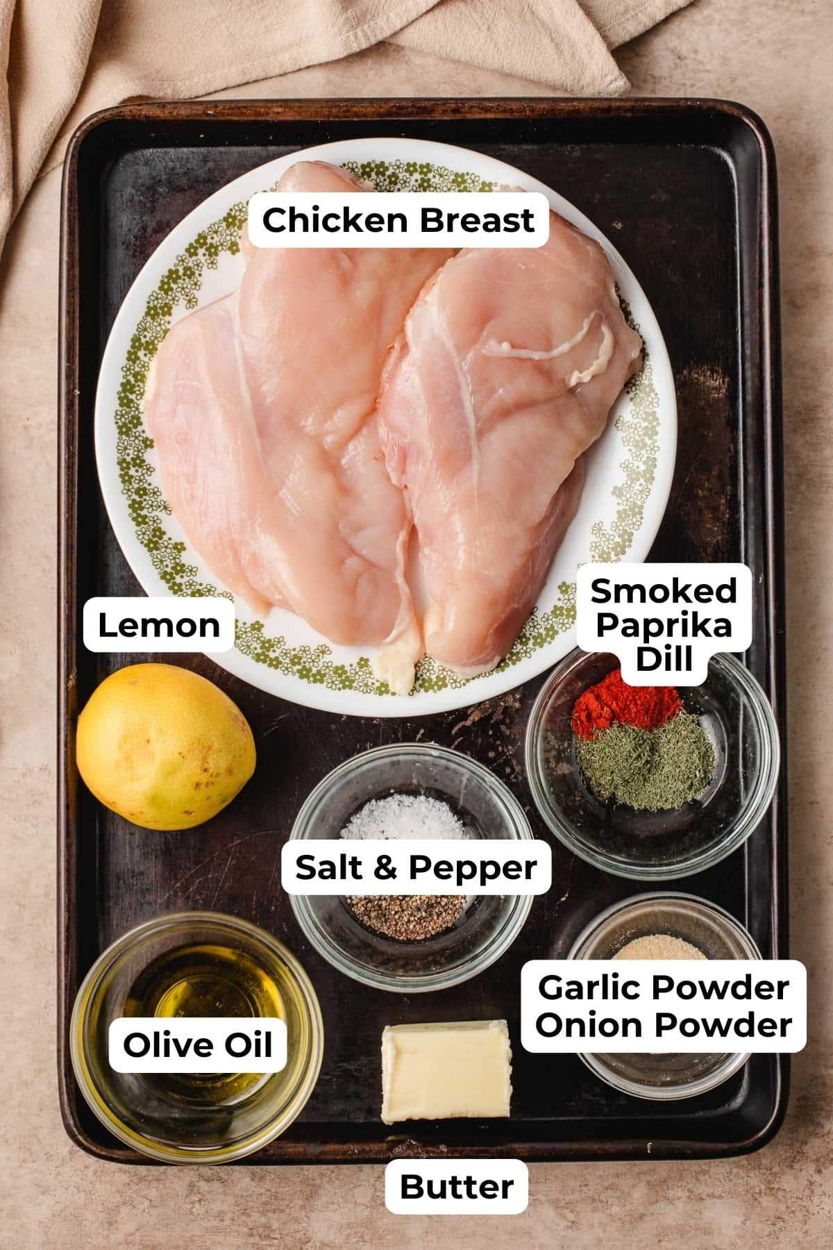 Raw chicken breasts on a plate, surrounded by a lemon, seasonings, olive oil, and butter.