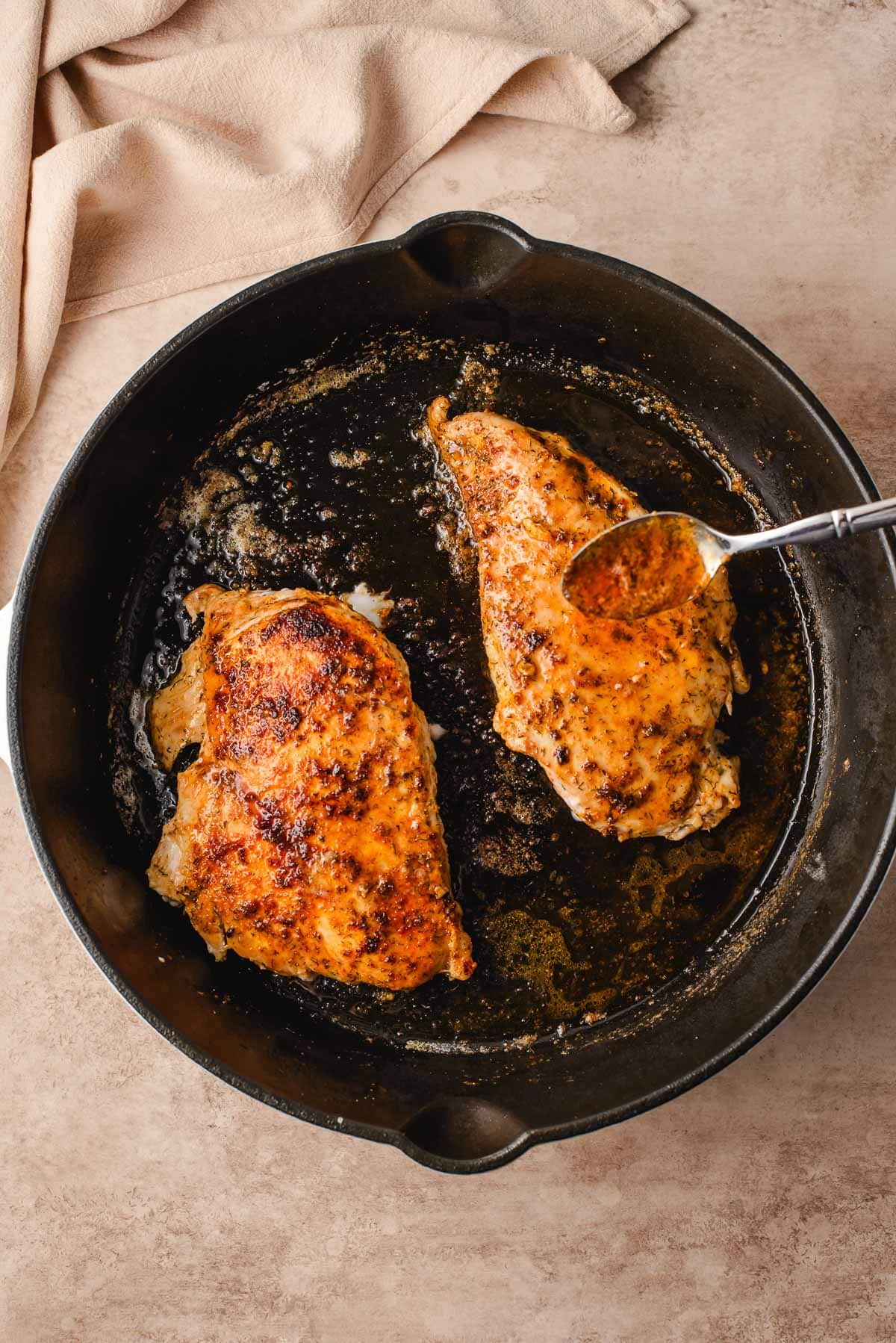 A spoon drizzling butter over chicken breasts in a cast iron skillet.